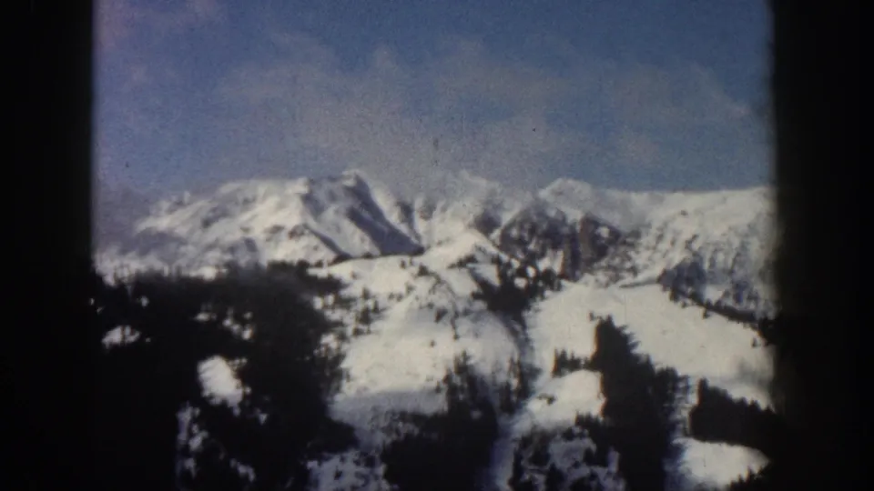 8mm home movie frame from Aspen, Colorado, 1961, showing snow-laden peaks and conifers under a pale blue winter sky with scattered clouds.