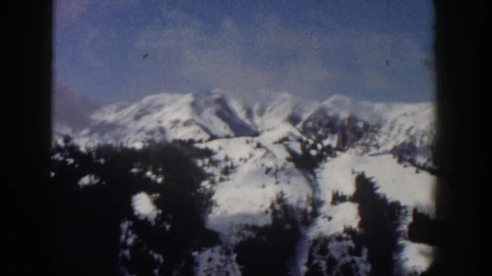 8mm home movie pan frame from Aspen, Colorado, 1961, showing a broader sweep of snow-covered mountains and conifer forest under blue sky.