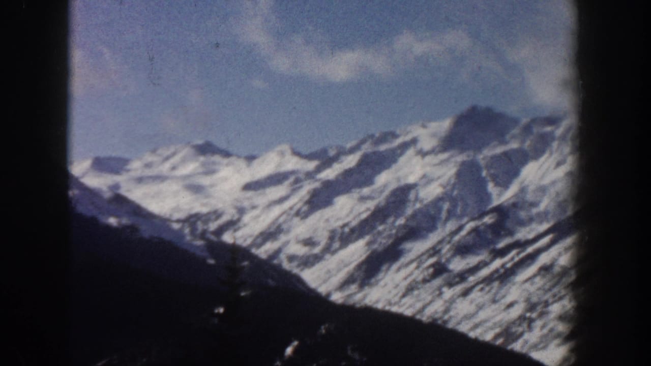 Snow-capped alpine peaks under blue sky with white clouds, dark window-frame edges visible, 8mm film grain.