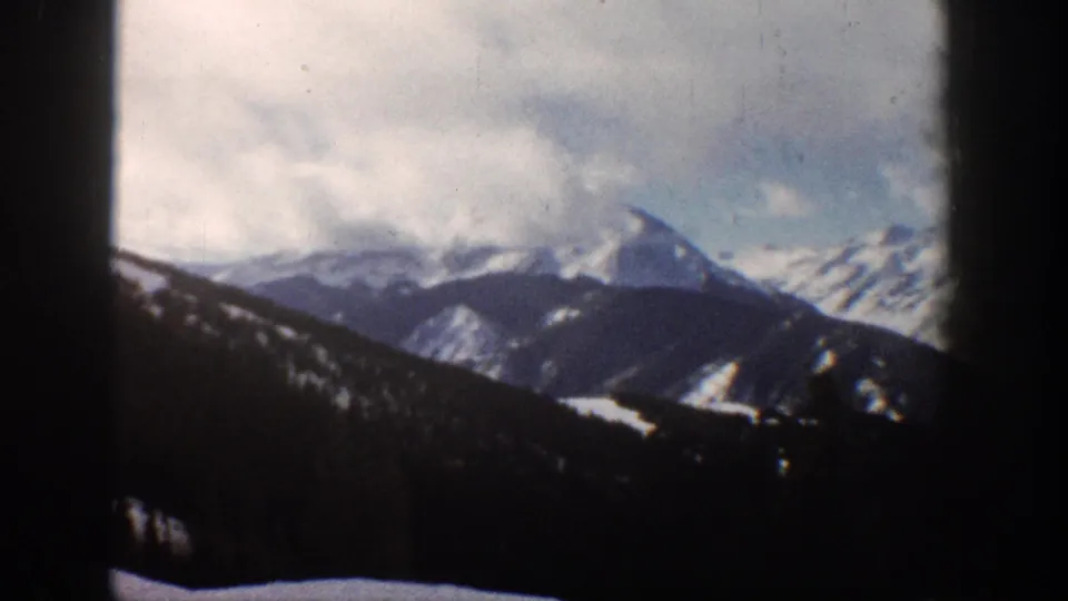 8mm home-movie frame from 1961 showing an Aspen-area mountain peak emerging from heavy cloud cover above a dark forested hillside.