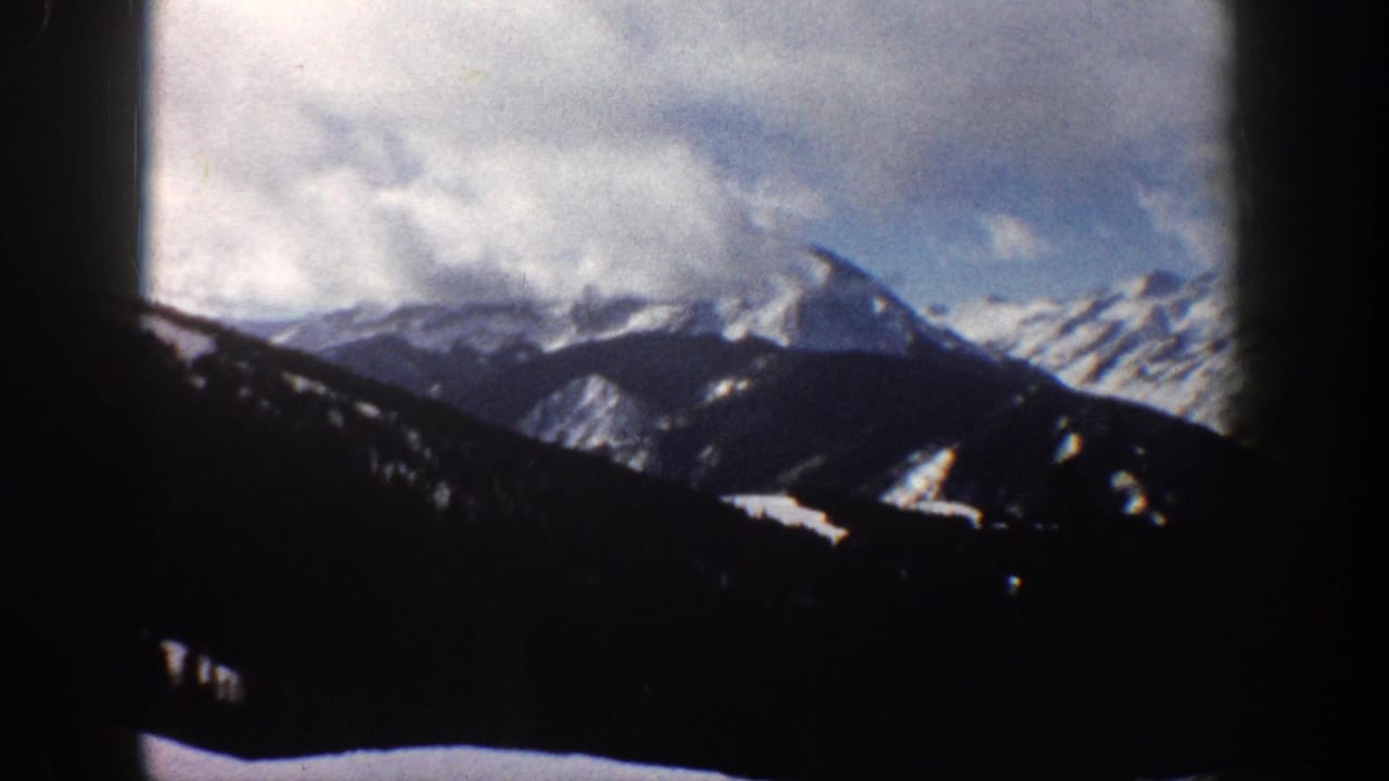 8mm view of snow-covered Rocky Mountain peaks near Aspen, Colorado, circa 1961, framed by a dark vehicle or window silhouette at left, dramatic clouds above.