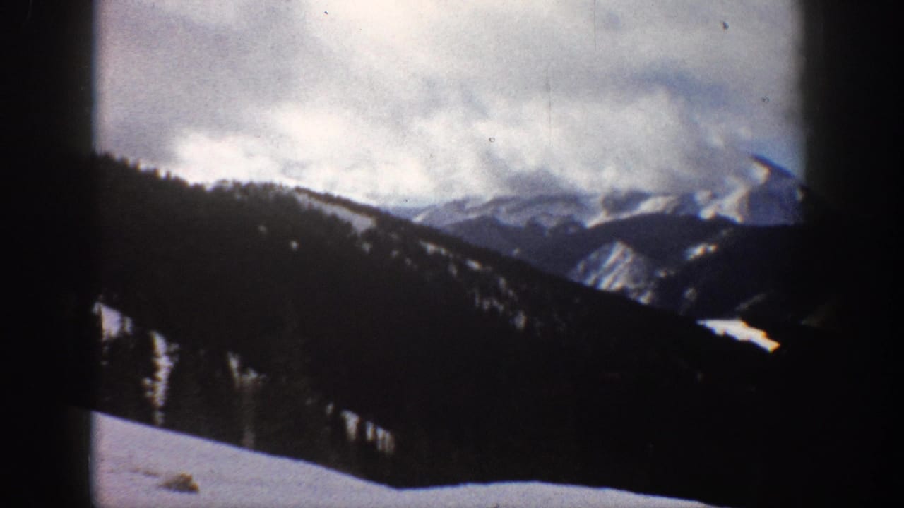 Mountain range with snow-covered peaks and conifer forest under cloud cover, framed by dark 8mm projector border