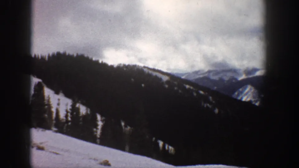 Colorado Rocky Mountain winter scene circa 1961, prominent conifers and snow slope with distant peaks under heavy cloud cover, 8mm color home movie