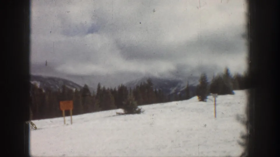 8mm home movie frame, Aspen Colorado area, 1961 — open snowfield with a rectangular wooden sign on a post and a second taller post, forested ridge beyond.