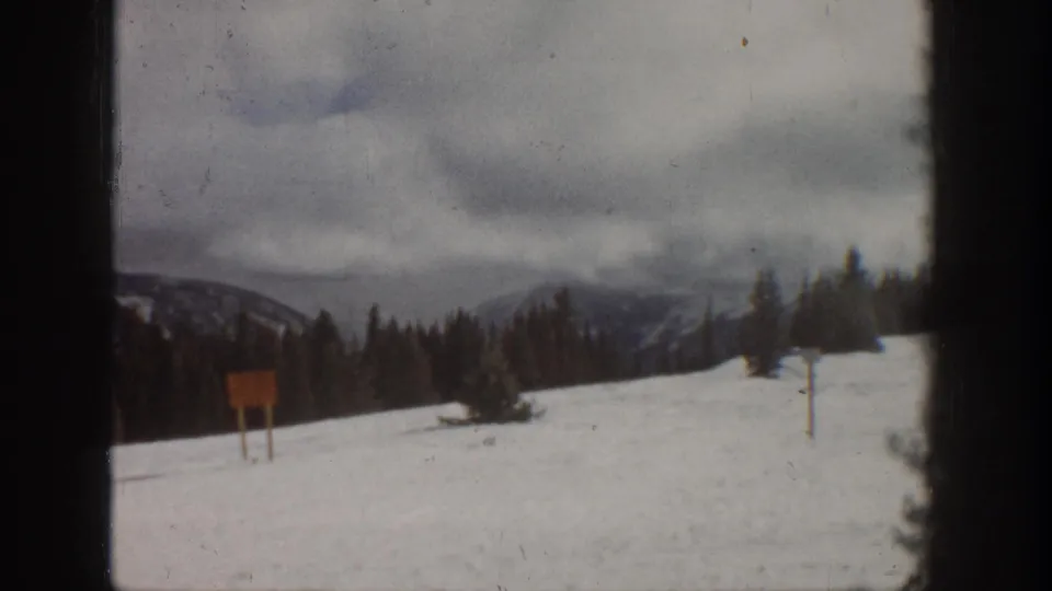 8mm home movie frame, Aspen Colorado area, 1961 — near-identical snowfield view with wooden signpost pair and cloudy mountain backdrop.