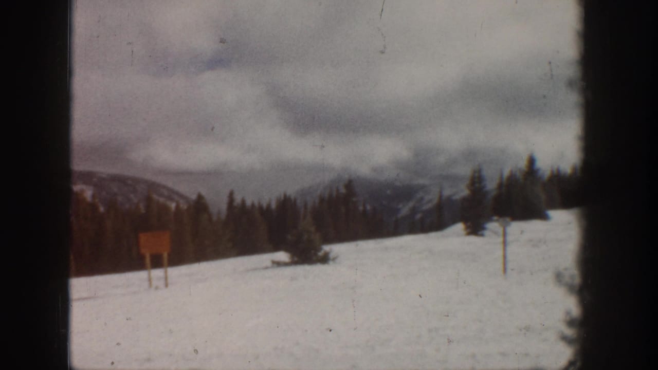 8mm home movie frame from what metadata identifies as Aspen, Colorado, circa 1961 — snow-covered slope, cloudy sky, evergreen treeline, and a wooden signboard on posts.