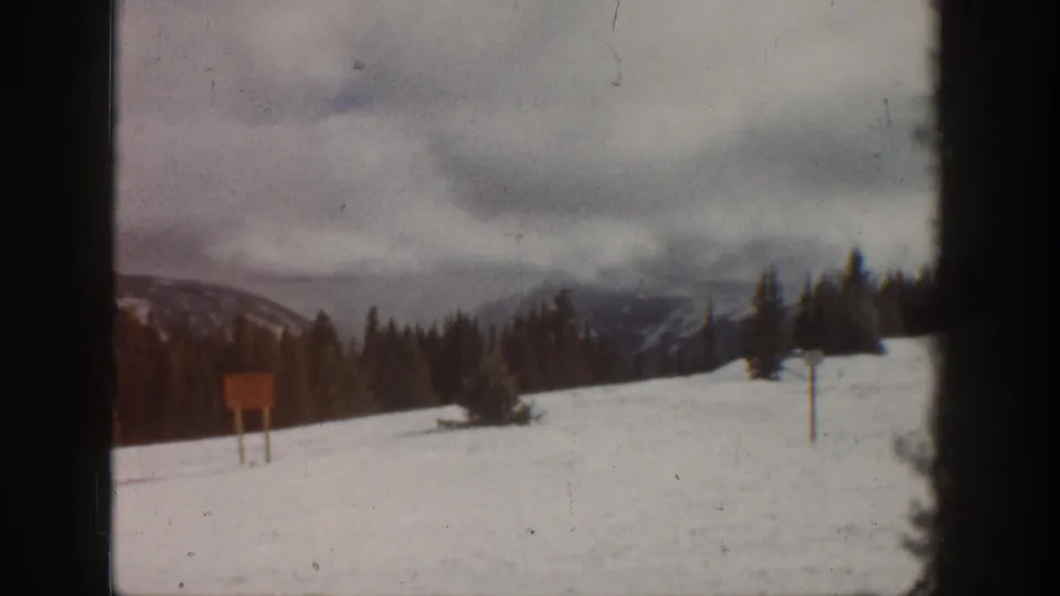 8mm home movie frame from what metadata identifies as Aspen, Colorado, circa 1961 — snow-covered slope, cloudy sky, evergreen treeline, and a wooden signboard on posts.