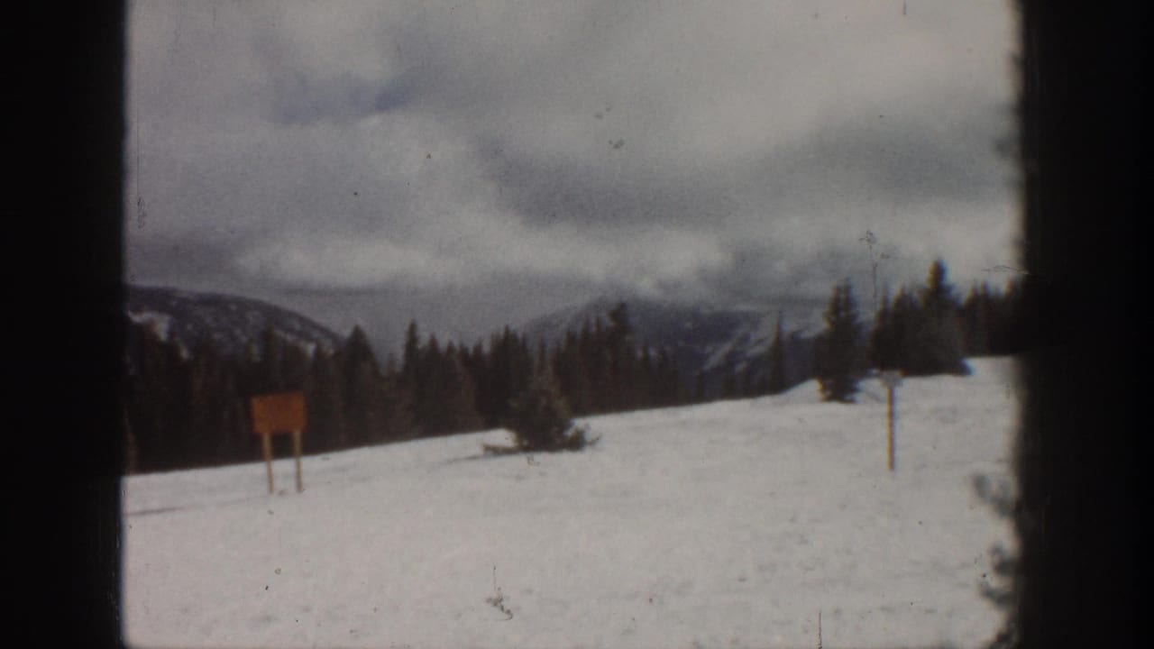 Second 8mm frame from the same ski slope scene — overcast sky, dense pine treeline, and a wooden signboard consistent with an early-1960s Colorado ski resort setting.