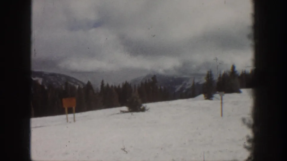 Second 8mm frame from the same ski slope scene — overcast sky, dense pine treeline, and a wooden signboard consistent with an early-1960s Colorado ski resort setting.