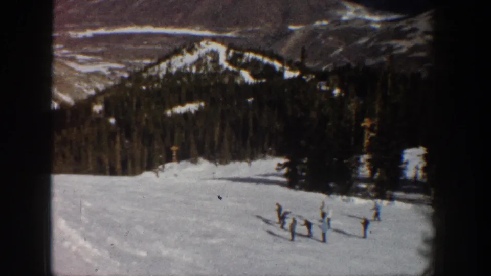 8mm home movie frame showing a snow-covered mountain valley at what metadata identifies as Aspen, Colorado, 1961 — small figures on the slope foreground with ski lift poles and distant ridgelines.