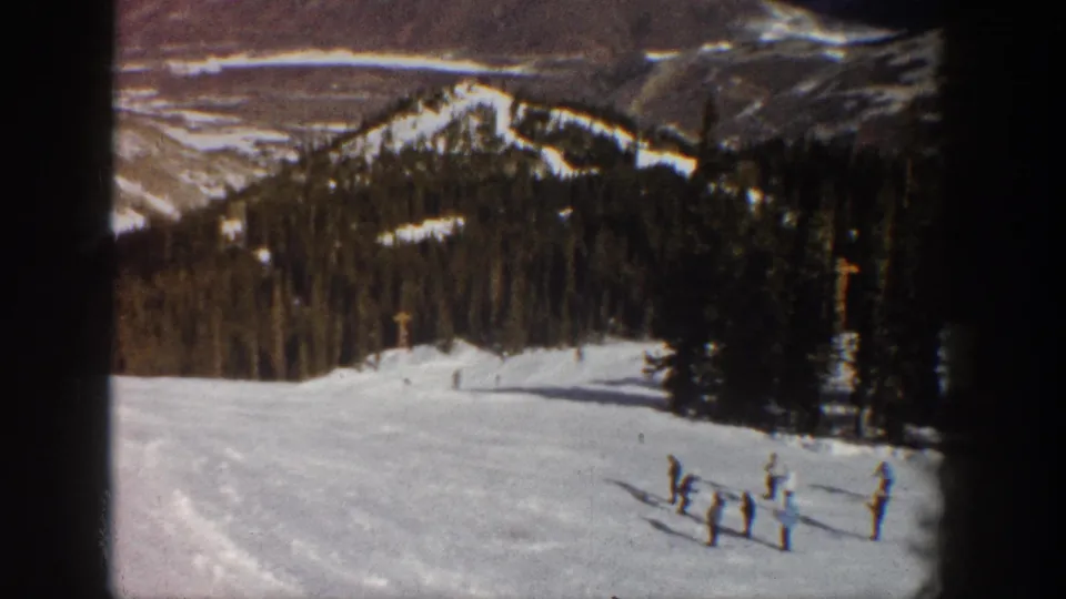Final 8mm frame showing a cluster of figures on a wide snowy slope, with snow-dusted Rocky Mountain peaks and dense treeline — consistent with Aspen, Colorado, circa 1961.