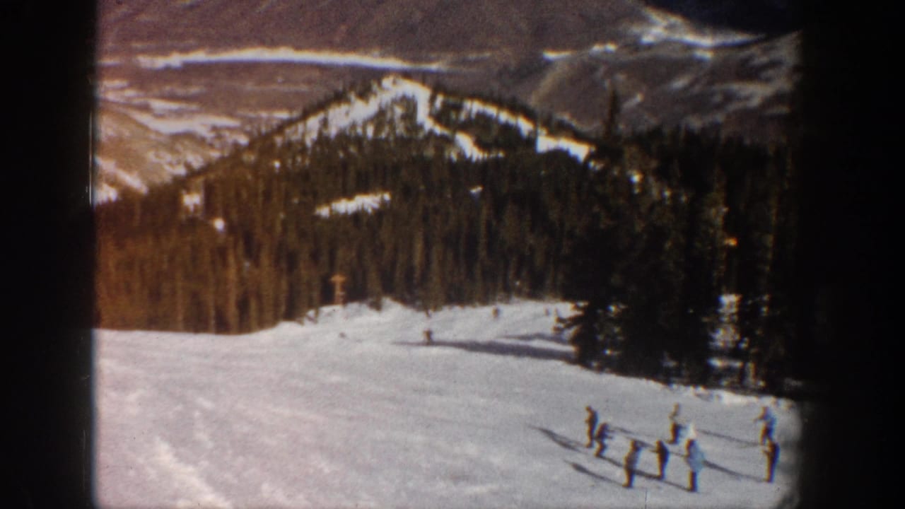 1961 8mm home movie frame showing skiers gathered on a sunny snow slope at what is described as Aspen, Colorado, with conifer forest and mountain peaks visible.