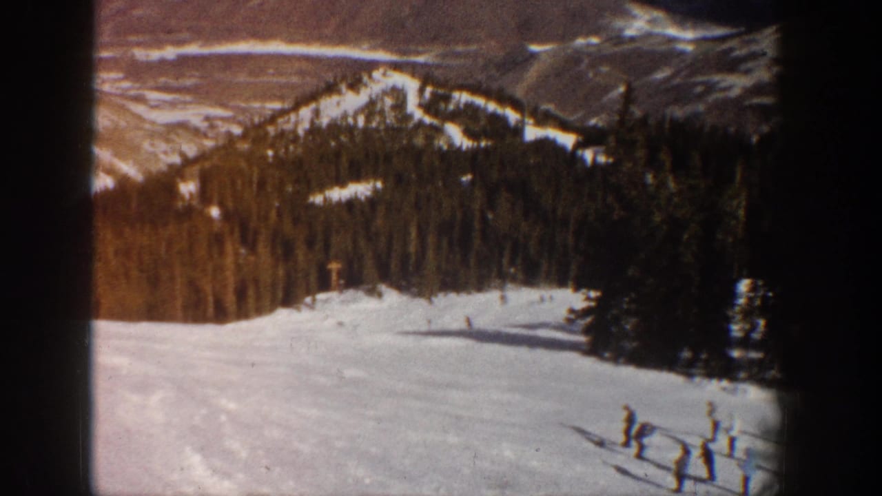 Continuation of 1961 8mm ski footage showing skiers on a sunny Aspen-area slope; conifer forest and bare mountain ridgeline remain visible in background.