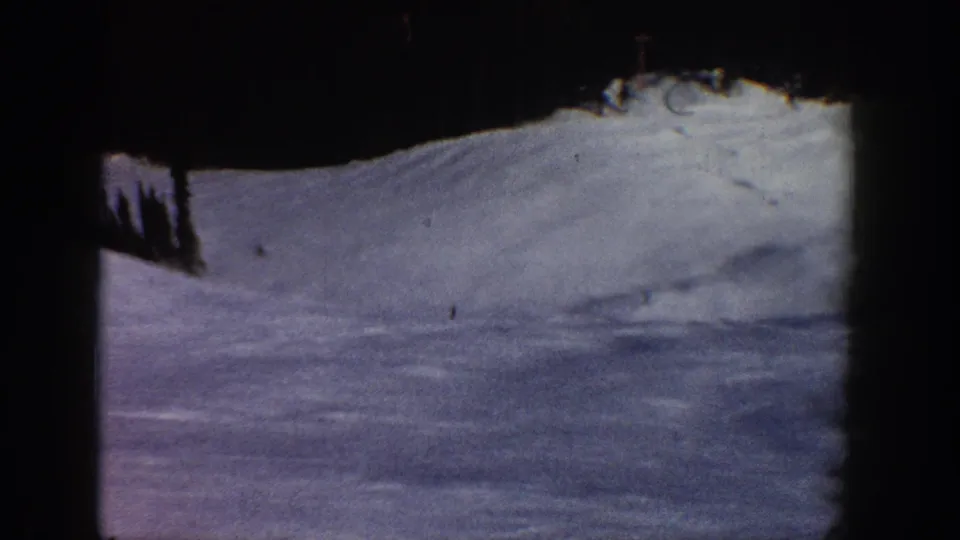 8mm home movie frame from Aspen area ski footage showing a shadowed snow slope with dense conifer canopy at frame top; cool blue-purple tones suggest shade or camera panning.