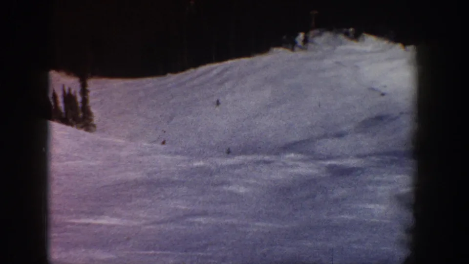 Wide ski slope view, likely Aspen, Colorado, circa 1961; 8mm color shift gives snow a blue-purple cast; tiny distant skiers visible; evergreen trees at ridge.