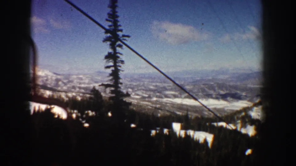 View from upper mountain showing ski lift cables, a tall evergreen tree, and a sweeping snow-covered valley; likely Aspen, Colorado, circa 1961, on 8mm film.