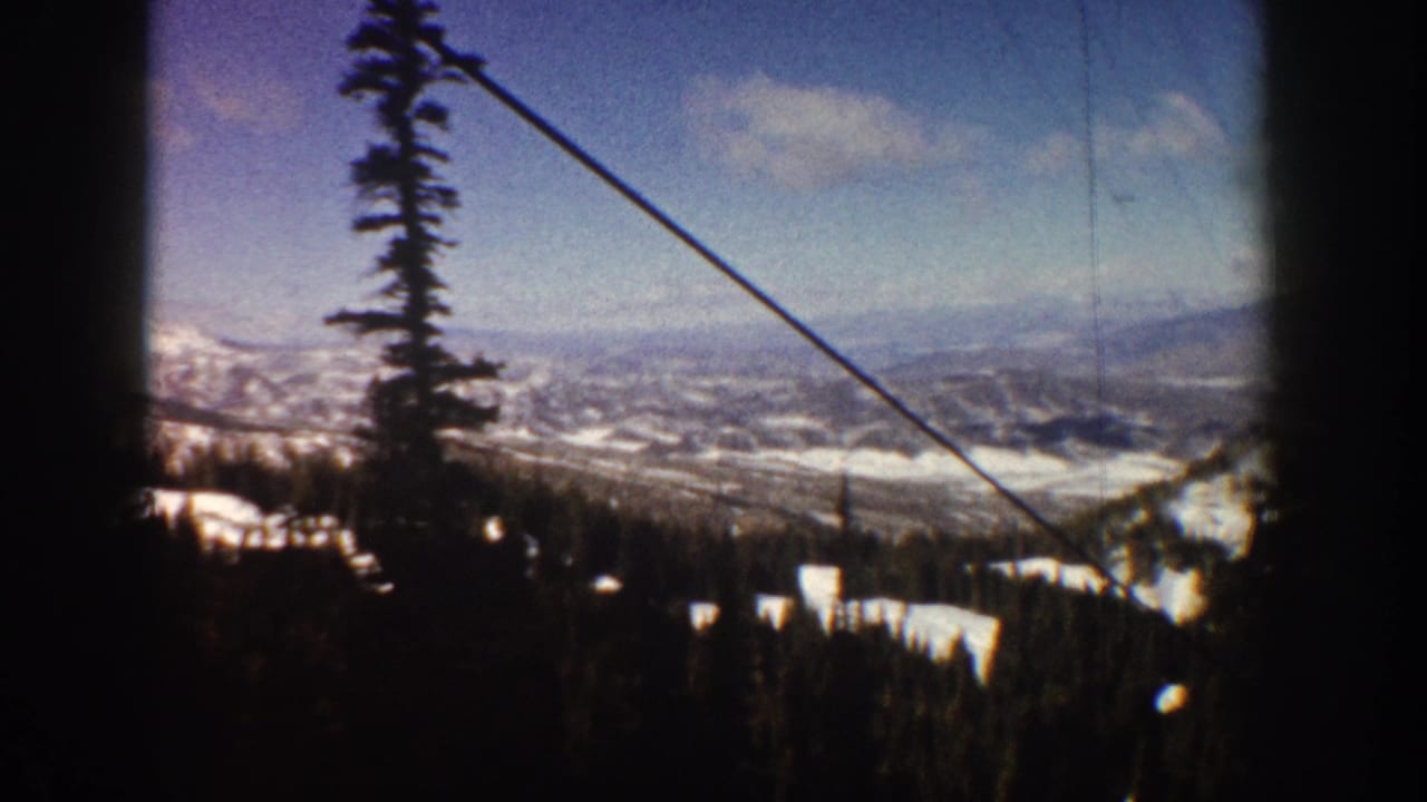 8mm home movie frame from a ski lift at Aspen, Colorado, circa 1961, showing snowy mountain slopes, dense conifer forest, and a distant valley below.