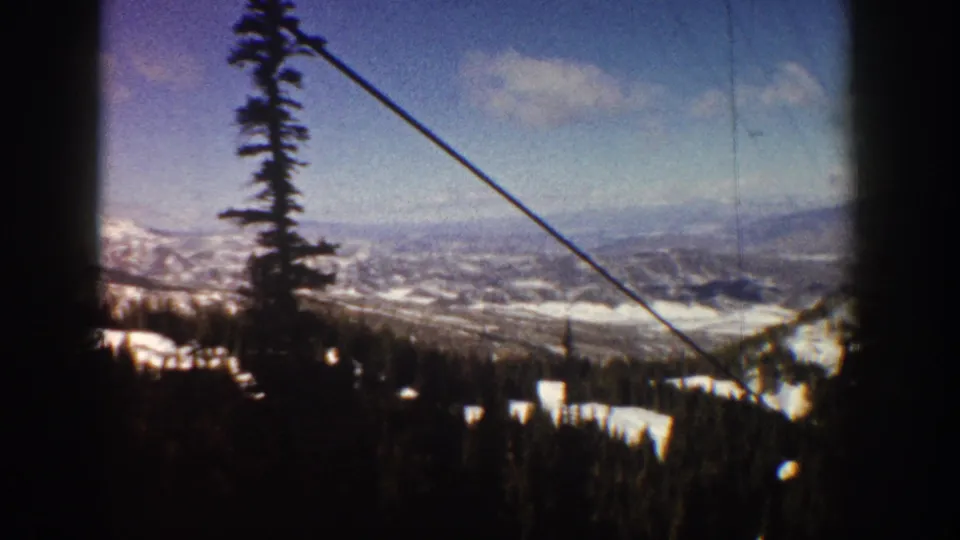 8mm home movie frame from a ski lift at Aspen, Colorado, circa 1961, showing snowy mountain slopes, dense conifer forest, and a distant valley below.