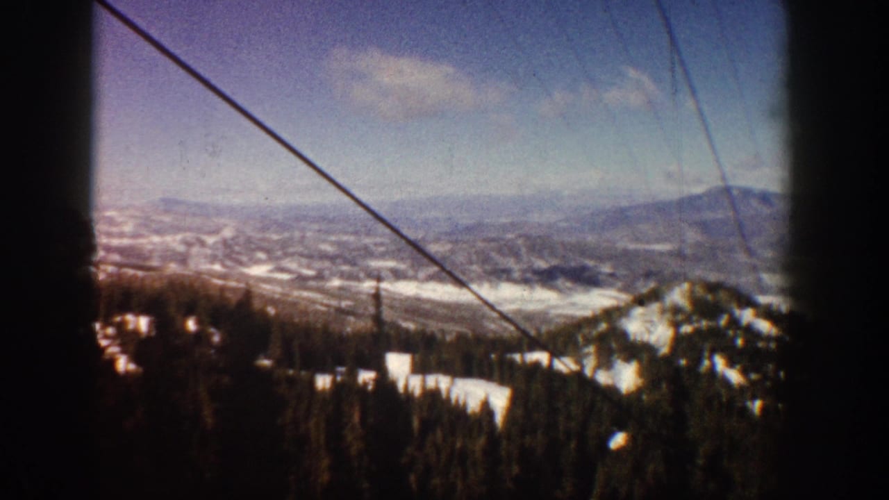 Ski lift cable view over a snow-dusted forested mountain ridge with a distant peak and valley below, filmed on 8mm.