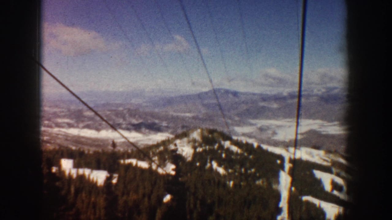 View from a ski lift over snow-patched mountain slopes and dense evergreen forest, lift cables crossing the upper frame.