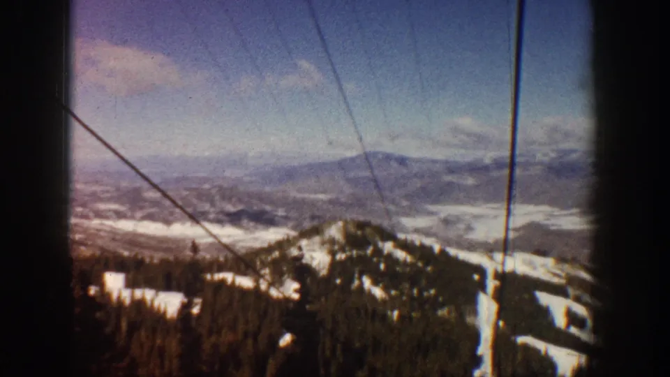 8mm home-movie frame, Aspen, Colorado, circa 1961; ski lift cables frame a downward view of snow-covered slopes, evergreen forest, and a wide mountain valley.