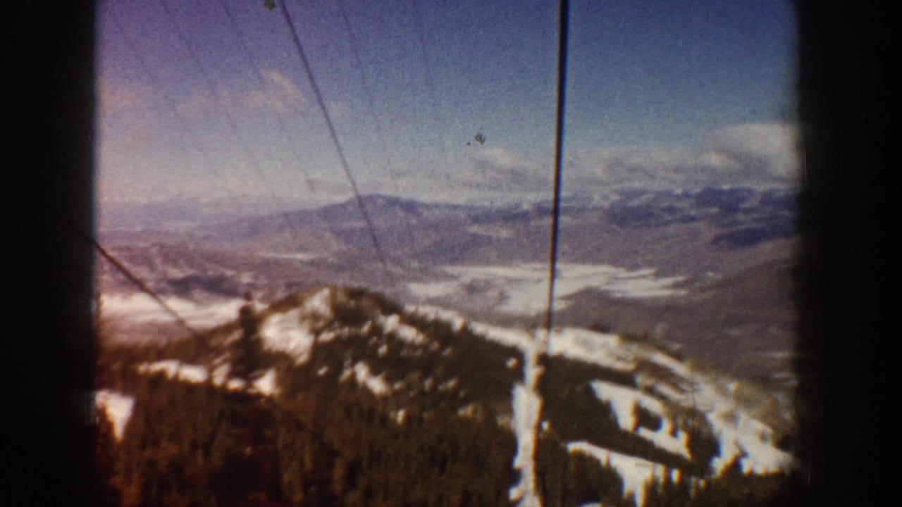8mm home-movie frame, Aspen, Colorado, circa 1961; lift cables cross the upper frame as the ride ascends past a snow-patched peak and a broad valley below.