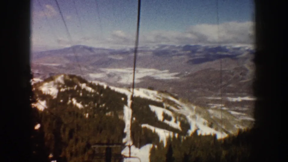 8mm home-movie frame, Aspen, Colorado, circa 1961; lift cables arc across the upper left as the camera captures sweeping mountain and valley scenery.