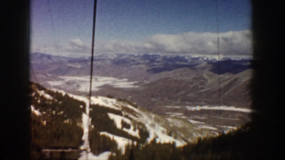 8mm home-movie frame, Aspen, Colorado, circa 1961; the ride continues upward with cables, snow-patched slopes, evergreen stands, and a wide valley receding below.
