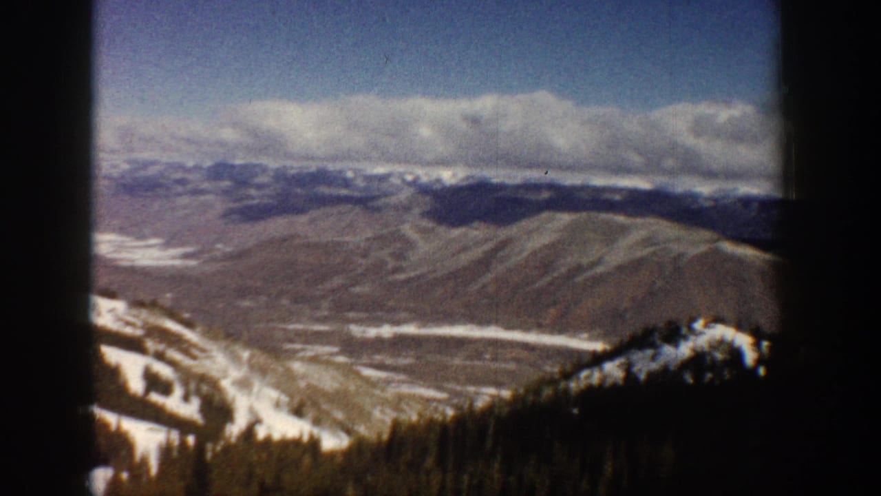 8mm panorama continuing a ski-lift traverse across a broad Colorado mountain valley with distant snow-capped peaks and forest belt, circa 1961.