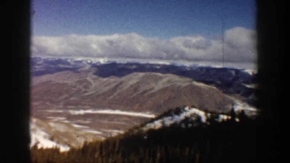 8mm ski-lift elevation shot of an expansive Colorado mountain valley; snow patches on ridges and evergreen forest visible below, circa 1961.