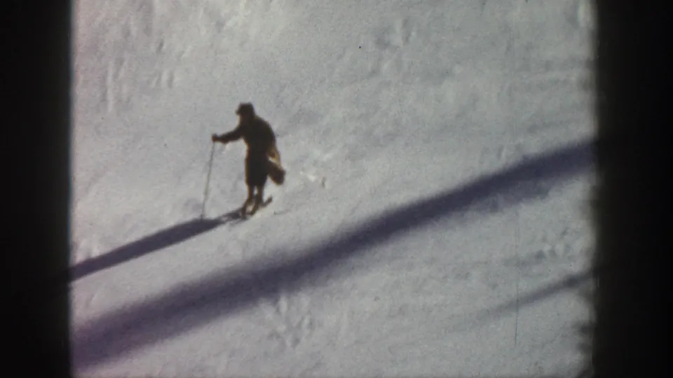 A single dark-clad skier descends a sunlit slope with ski poles, silhouetted against white snow with strong diagonal shadow lines; Aspen, Colorado, circa 1961.