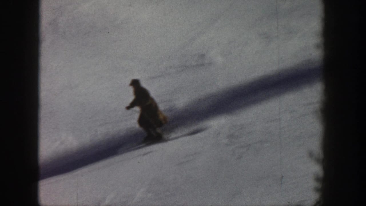 8mm home movie frame, circa 1961, Aspen Colorado — lone skier descending open snow slope, long diagonal shadow across white terrain.