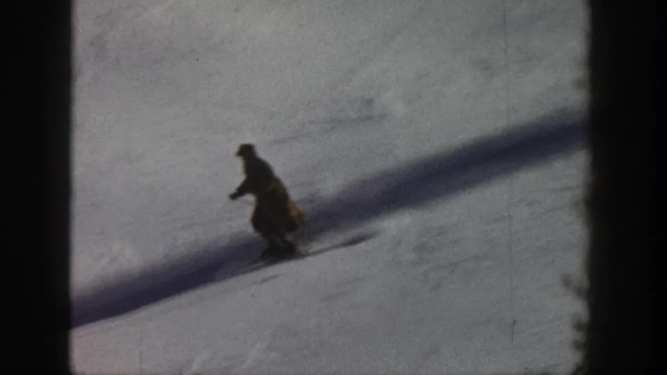 8mm home movie frame, circa 1961, Aspen Colorado — lone skier descending open snow slope, long diagonal shadow across white terrain.