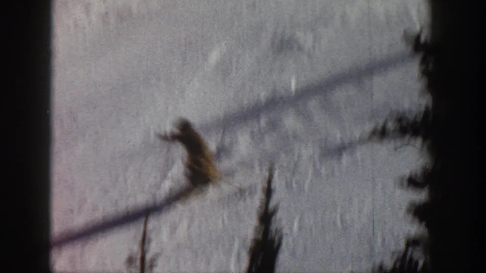 8mm home movie circa 1961, Aspen Colorado — skier mid-descent on open slope, tree line at right, warm color shift from film aging.