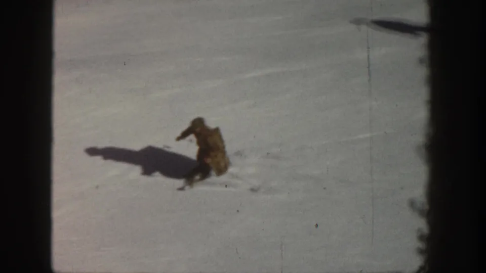 8mm home movie circa 1961, Aspen Colorado — skier crouched mid-turn on open slope, long shadow visible, trees at frame edge.