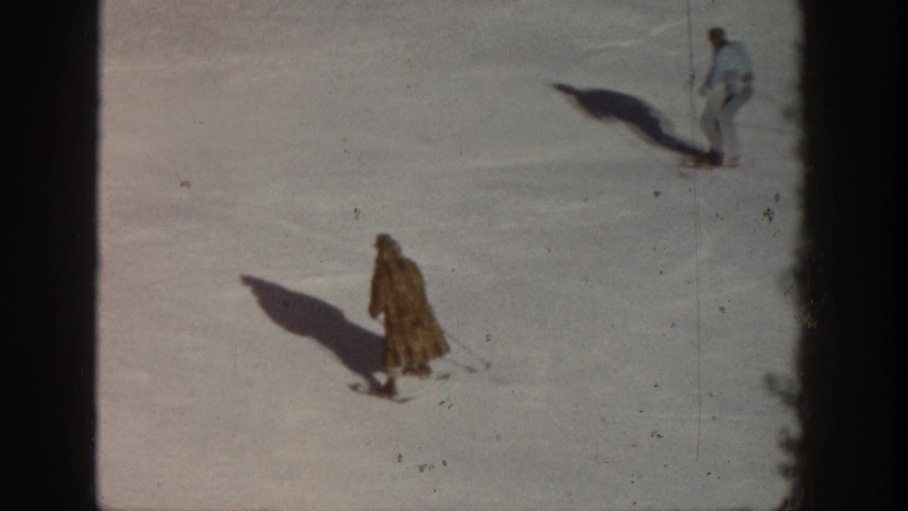 Two figures on an open sunlit snow slope; the foreground figure wears a long tan coat and casts a dramatic shadow; a second figure stands further up the hill.