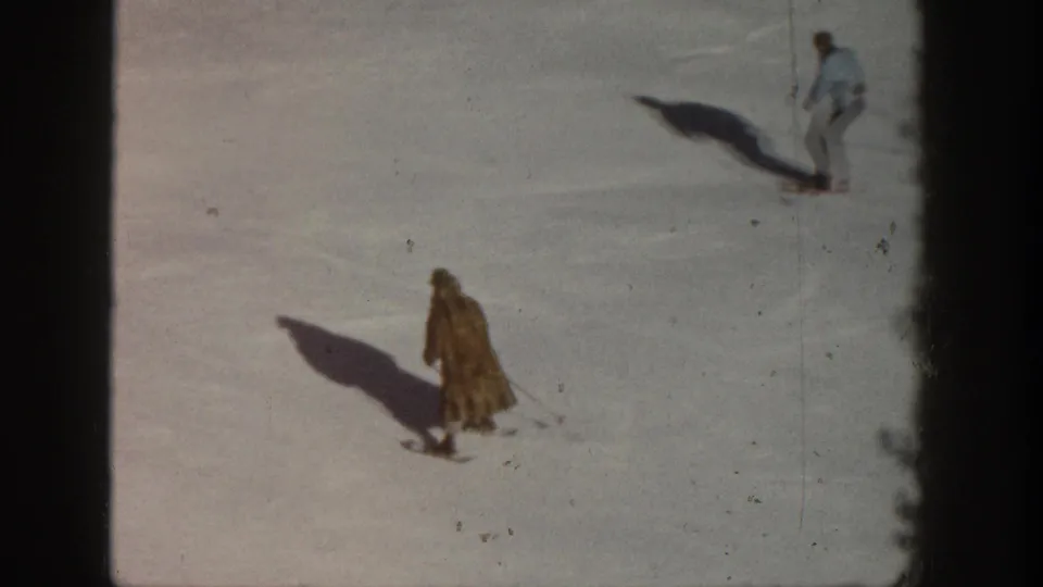 Two skiers on a sunlit open snow slope in what is identified as Aspen, Colorado, circa 1961; the foreground figure wears a long tan coat with both casting long shadows across the snow.