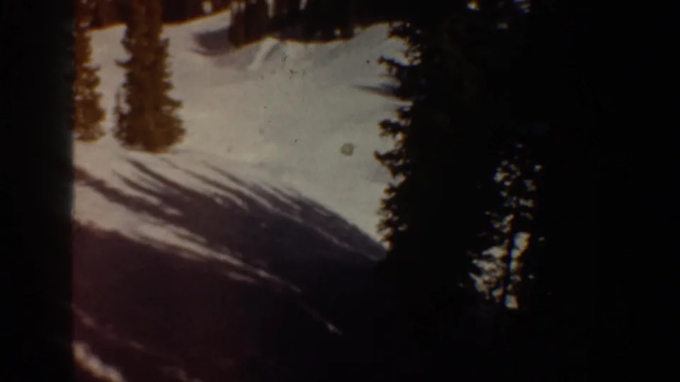 Snow-covered ski slope with conifer trees and sweeping diagonal tree shadows, in what is identified as Aspen, Colorado, circa 1961, shot on 8mm film.