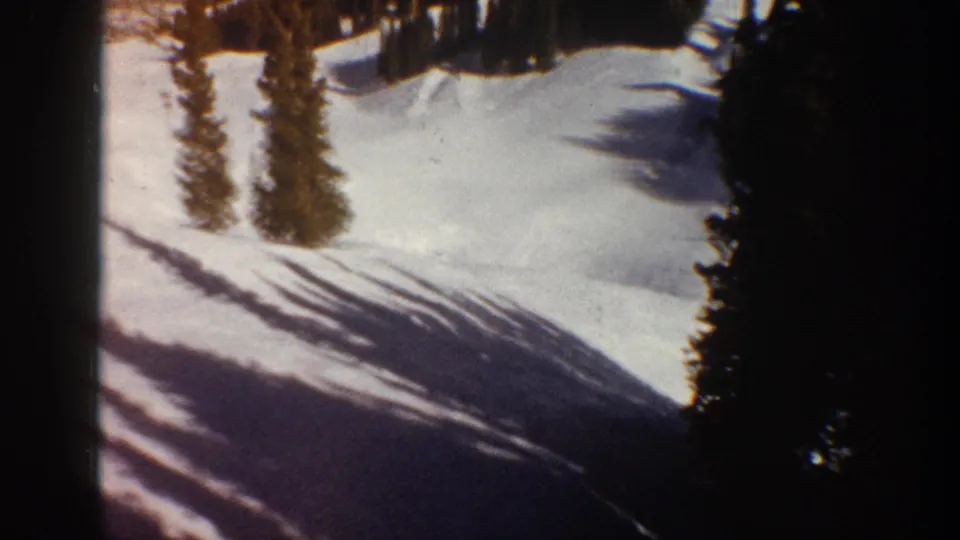 Snow-covered ski slope lined with dense conifers and long diagonal tree shadows in what is identified as Aspen, Colorado, circa 1961, shot on 8mm.