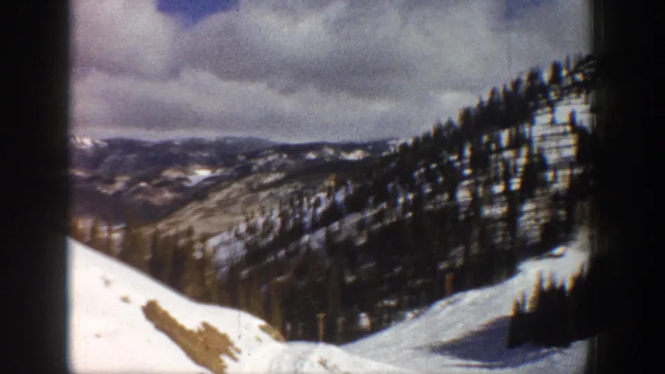 Panoramic view of snow-covered Colorado mountain ridgelines with dense conifer forest under overcast sky, circa 1961, 8mm home movie.