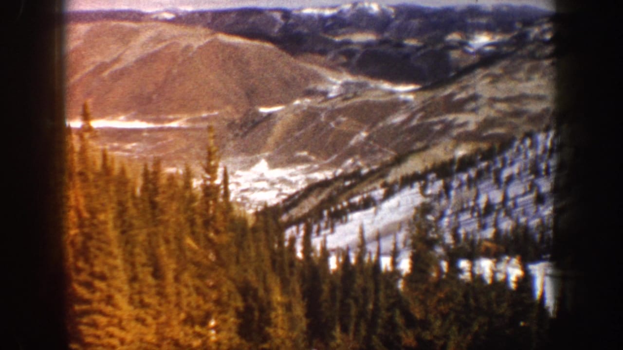 Mountain valley with snow patches, pines, and hills beyond a dark frame edge.