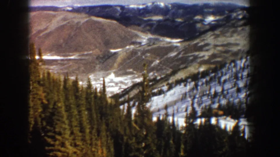 1961 Aspen, Colorado home-movie frame showing pines, patchy snow, brown slopes, and distant mountain ridges.