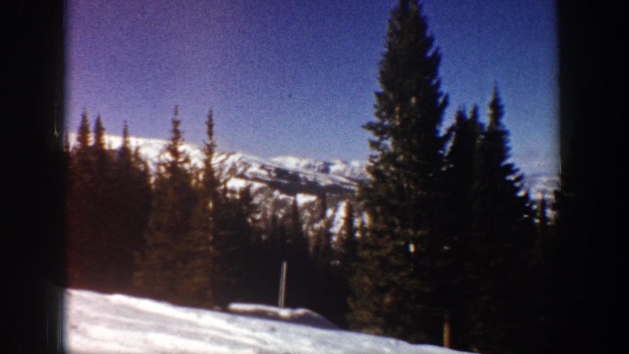 1961 Aspen winter view with evergreen trees in the foreground, distant snow-covered ridges, blue sky, and a small post near the snow.