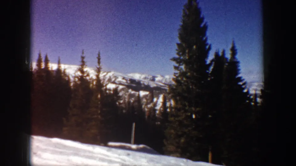 1961 Aspen winter view with evergreen trees in the foreground, distant snow-covered ridges, blue sky, and a small post near the snow.