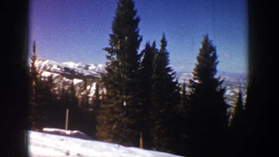 1961 Aspen winter landscape with tall evergreens, snow-covered ridges, blue sky, and distant hazy terrain visible through the trees.