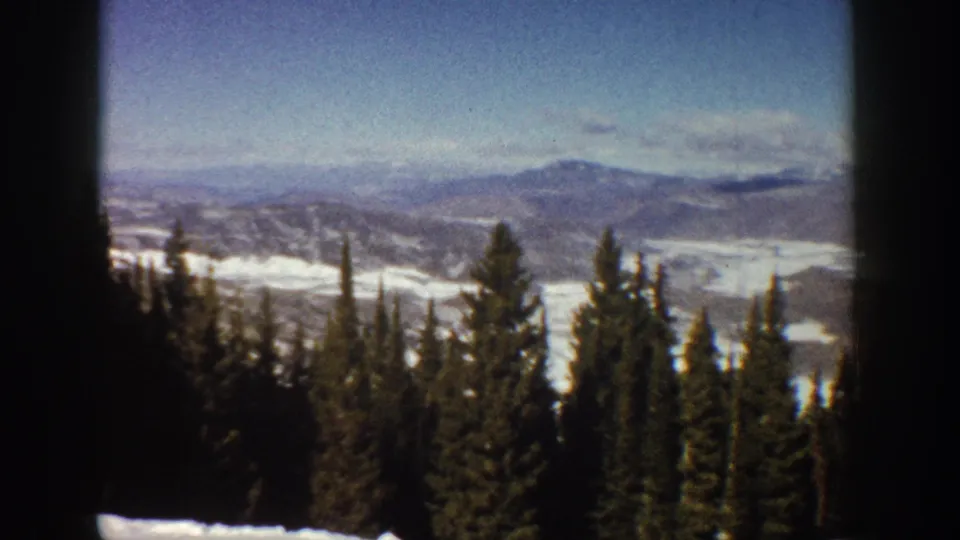 1961 Aspen-area 8mm frame showing dark pines below a broad winter valley, with blue-gray mountains and soft clouds on the horizon.