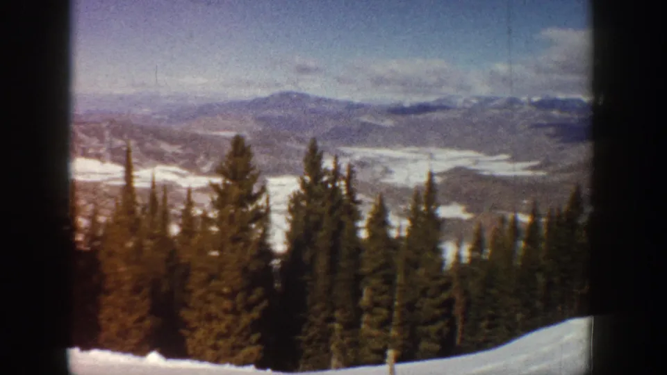 1961 Aspen-area 8mm view with a snowbank at the lower edge, evergreen pines, scattered snow, and mountain ridges in the distance.