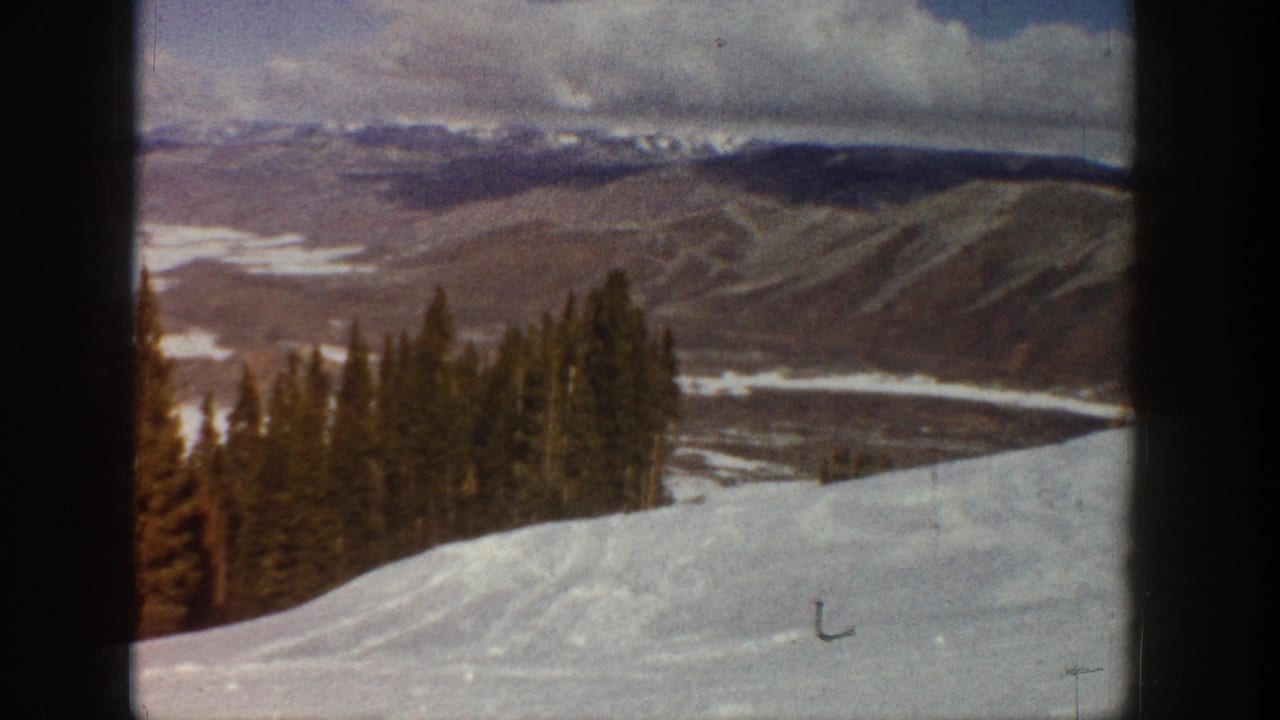 1961 Aspen-area 8mm frame showing a snow-covered slope, dark evergreens, distant mountain ridges, and a cloudy winter sky.