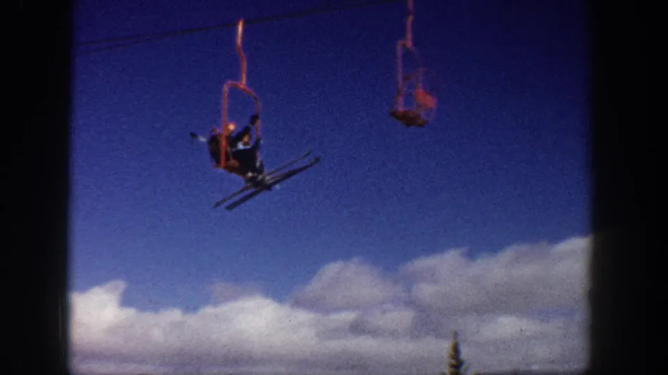 1961 Aspen-area ski lift shot with distant skiers seated on chairs crossing a blue sky above clouds and treetops.