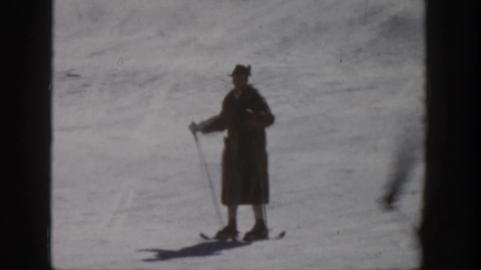 1961 Aspen home-movie still of an adult skier in a long coat and brimmed hat standing with poles on the snow.